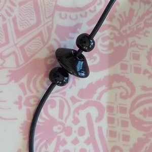May include: A black necklace featuring a thin, flexible cord strung with three distinct black beads. The central bead is shaped like a flattened disc, flanked by two smaller, spherical black beads, all displaying a smooth, glossy finish.