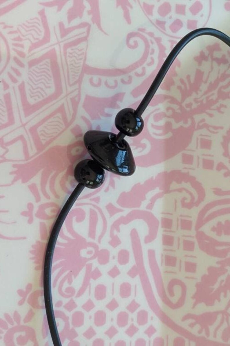 May include: A black necklace featuring a thin, flexible cord strung with three distinct black beads. The central bead is shaped like a flattened disc, flanked by two smaller, spherical black beads, all displaying a smooth, glossy finish.