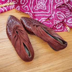 In this photo, the mules are depicted to show the ruched, draped leather at the cuff. These mules are croc embossed, leather in a shade of golden russet brown. The shoes are displayed on a wooden plank surface, with a chiffon scarf of pink and white geometric patterns.