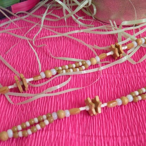 K&ouml;nnte beinhalten: Eine Holzperlenkette mit braunen und wei&szlig;en Perlen und sternf&ouml;rmigen Holzanh&auml;ngern. Die Kette liegt auf einer rosa strukturierten Oberfl&auml;che.