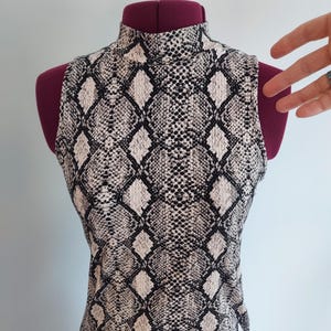 May include: Sleeveless mock neck top with a snakeskin pattern in black, white, and grey. The top is displayed on a maroon dress form. The snakeskin print is a repeating diamond pattern.