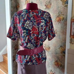 May include: A short-sleeved blouse featuring a vibrant, multi-colored mosaic-style pattern in shades of red, blue, white, and black. The garment is cinched at the waist with a wide, solid magenta-colored belt that features a rectangular buckle.