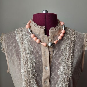May include: A beige lace top with a pink and silver beaded necklace. The top has a high neckline and a button-up front.