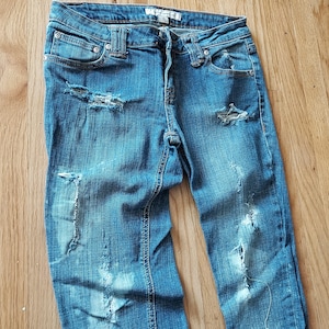 May include: Blue denim jeans with distressed details, including ripped sections on the front of the legs. The jeans have a classic five-pocket design and a button and zipper closure.