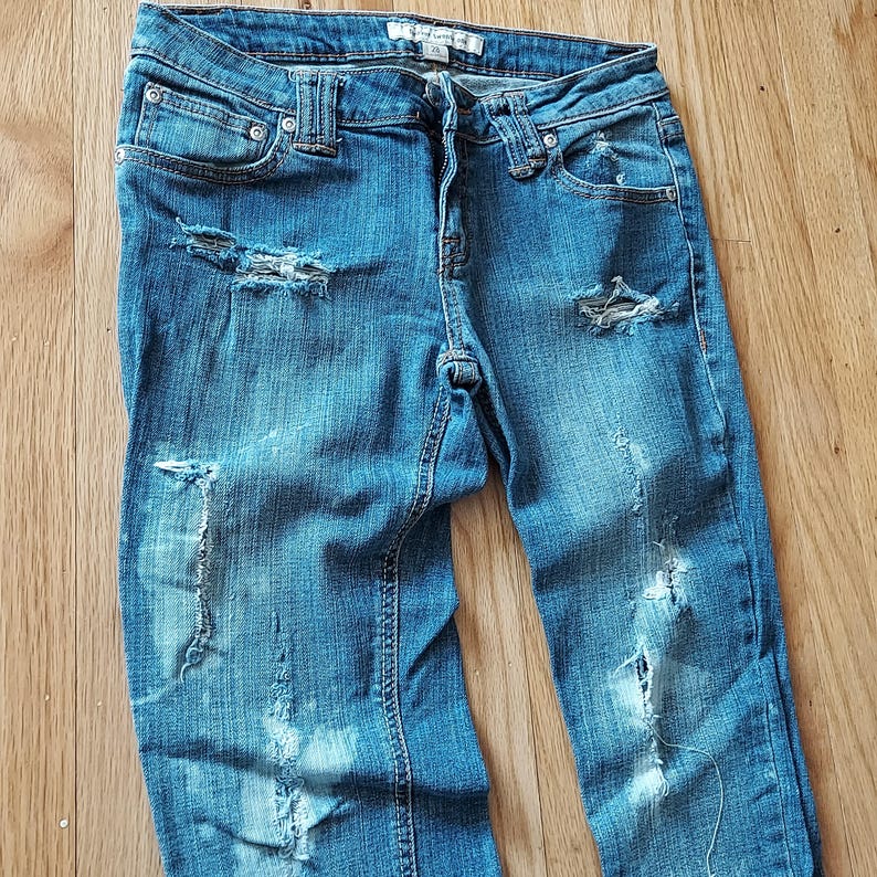 May include: Blue denim jeans with distressed details, including ripped sections on the front of the legs. The jeans have a classic five-pocket design and a button and zipper closure.
