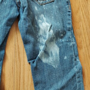 May include: Close-up of blue denim jeans with white bleach spots. The jeans have a classic five-pocket design and are made of a durable denim fabric. The jeans are on a wooden surface.