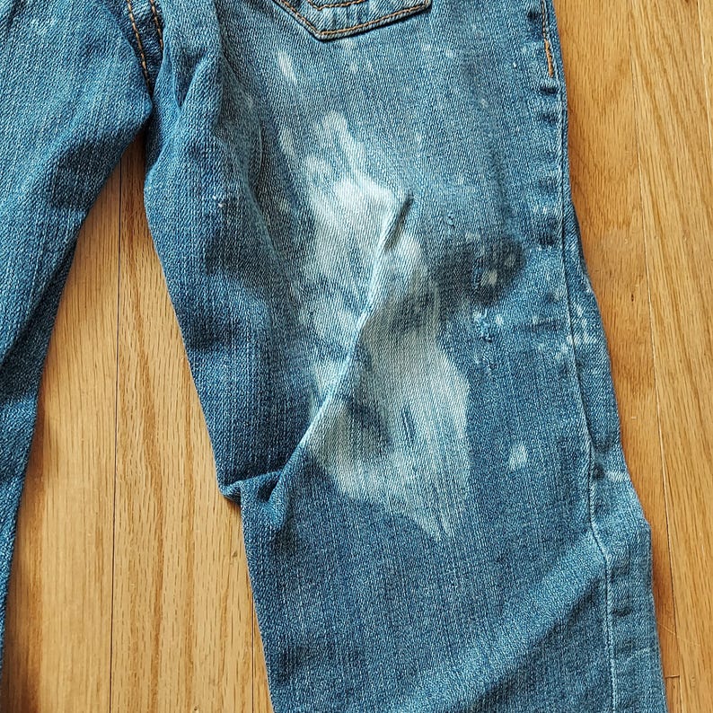 May include: Close-up of blue denim jeans with white bleach spots. The jeans have a classic five-pocket design and are made of a durable denim fabric. The jeans are on a wooden surface.