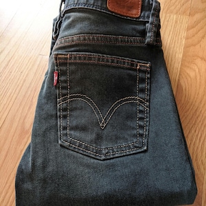 May include: A pair of dark blue denim jeans with a brown leather patch and red stitching. The jeans are folded in half and the back pocket is visible.