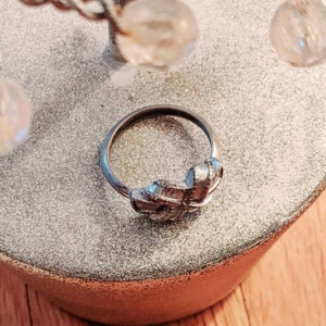 May include: A silver-toned metal ring featuring a textured, knot-like design at the center. The band is thin and circular, with a brushed metallic finish on the decorative knot element that creates a subtle, layered appearance on the surface.