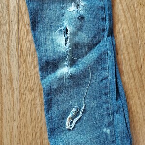 May include: Close-up of distressed blue denim jeans with a large tear and frayed edges. A long, loose thread hangs from the tear, creating a worn and vintage aesthetic. The jeans are on a wooden surface.