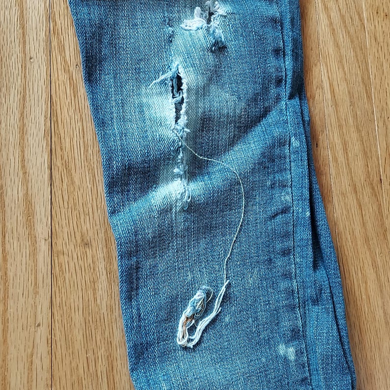 May include: Close-up of distressed blue denim jeans with a large tear and frayed edges. A long, loose thread hangs from the tear, creating a worn and vintage aesthetic. The jeans are on a wooden surface.