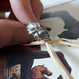 May include: A silver-toned ring featuring a textured, knotted ribbon design with a cross-hatched pattern across the surface. The metal has a brushed finish, creating a dimensional look with multiple loops that form a central knot structure.