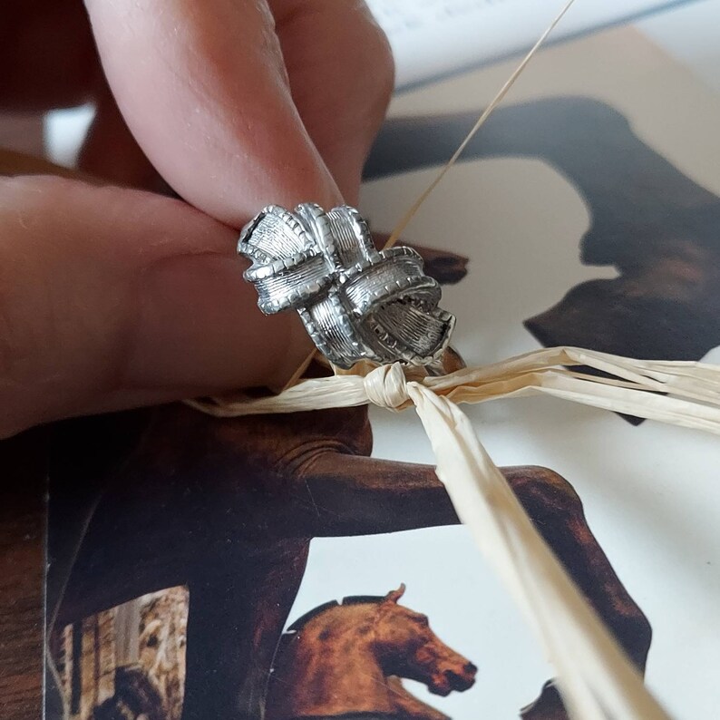 May include: A silver-toned ring featuring a textured, knotted ribbon design with a cross-hatched pattern across the surface. The metal has a brushed finish, creating a dimensional look with multiple loops that form a central knot structure.