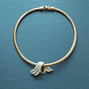 May include: A gold snake chain necklace with a white enamel pendant featuring a stylized wave design.