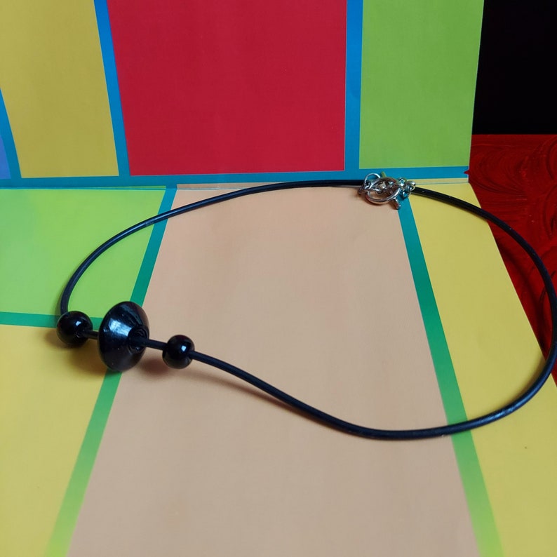 May include: A black cord necklace featuring three black beads, including one larger central disc-shaped bead flanked by two smaller spherical beads. The necklace is secured with a silver-toned metal toggle clasp at the back of the cord.