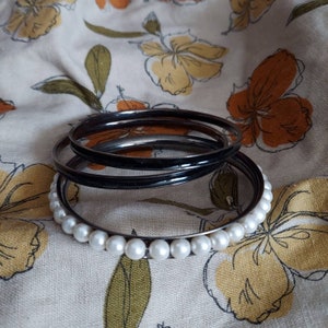 May include: A set of black bangle bracelets with a pearl accent. The bracelets are stacked on top of each other and are lying on a floral fabric.