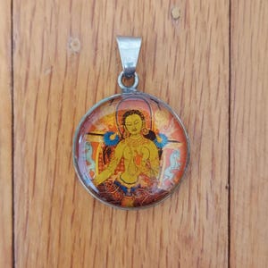 May include: Round pendant with a silver bail. The pendant features a colorful illustration of a deity with yellow skin, blue accents, and a red and orange background. The pendant is likely made of metal and glass.