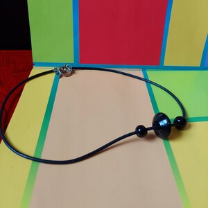 May include: A black cord necklace featuring a silver-toned toggle clasp and three black beads. The central bead is larger and disc-shaped, flanked by two smaller spherical beads on either side, all strung along a smooth, thin black cord.