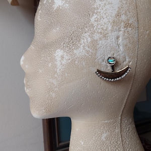 May include: A bronze-toned earring with a crescent shape, adorned with small, clear crystals along the curve. The earring features a small, round iridescent stone at the top. The earring is attached to a post.
