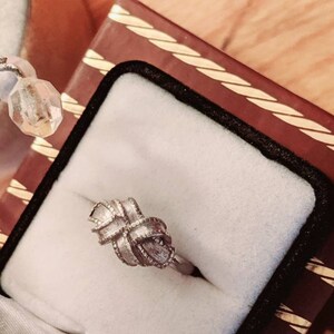May include: A silver-toned ring featuring a central knot design with a textured, ribbed surface. The band is smooth and circular, showcasing a detailed, woven-like pattern that forms a decorative knot shape at the top of the jewelry piece.