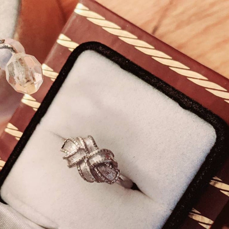 May include: A silver-toned ring featuring a central knot design with a textured, ribbed surface. The band is smooth and circular, showcasing a detailed, woven-like pattern that forms a decorative knot shape at the top of the jewelry piece.