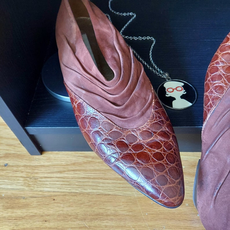 A pair of brown leather shoes with a pointed almond toe and a ruched suede cuff. The shoes have a crocodile-embossed pattern on the leather, and leather insoles like a moccasin. Displayed on a black wooden box, with a round pendant of red, black and white.