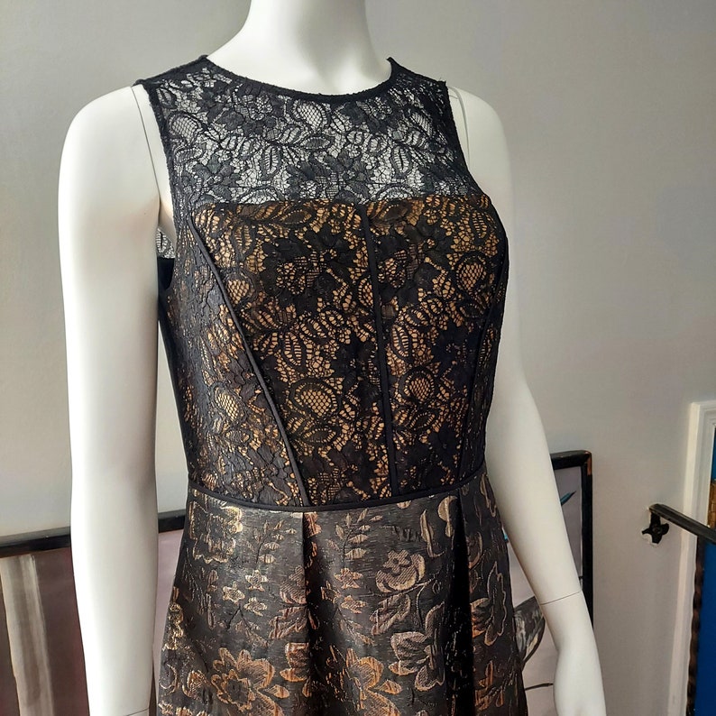 May include: A sleeveless black and gold dress on a mannequin. The bodice features black lace over a gold fabric, with a fitted design. The skirt is a darker gold with a floral pattern. The dress is designed for formal occasions.