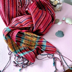 May include: A vibrant, handwoven scarf with a mix of red, purple, and teal hues. The scarf features striped and plaid patterns, with delicate white stitching and decorative tassels at the ends. The scarf is made of a soft, lightweight fabric.