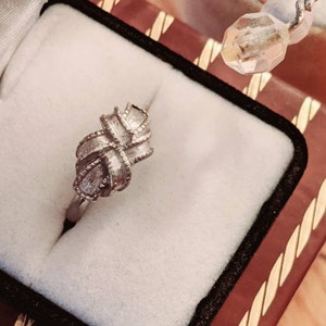 May include: A silver-toned ring featuring a textured, knot-like design with a central cross-over pattern. The ring is set within a white, velvet-lined jewelry box that has a dark brown exterior with gold-colored trim along the edges.