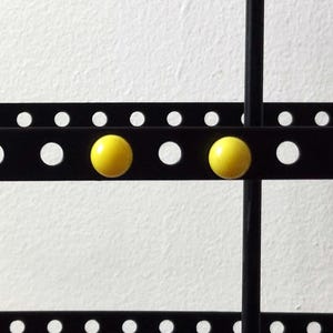 Puede incluir: Un expositor de pendientes de metal negro con múltiples barras horizontales y soportes verticales. El expositor presenta agujeros espaciados uniformemente y dos pendientes esféricos amarillos. El fondo es una pared blanca lisa.