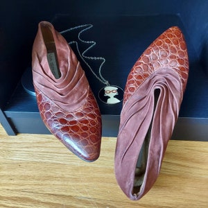 Rockabilly mules are made of Italian leather, in a croc embossed design. The slip-on style mules feature a cuff of ruched or draped suede, and a golden russet brown hue. The toes are pointed almond, and the heels are low stacked. Displayed on a black wooden box, with a round silver pendant which has a women's face wearing red eyeglasses.