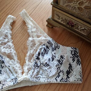 May include: A white and black snakeskin print bra with white lace trim. The bra is made of a soft material and has a classic design. The bra is a lingerie item.