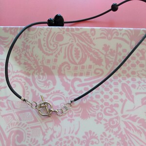 May include: A black cord necklace featuring a silver-toned toggle clasp with a circular ring and a T-bar. The cord is accented with two smooth, round black beads positioned along the length of the necklace, providing a simple and functional design.