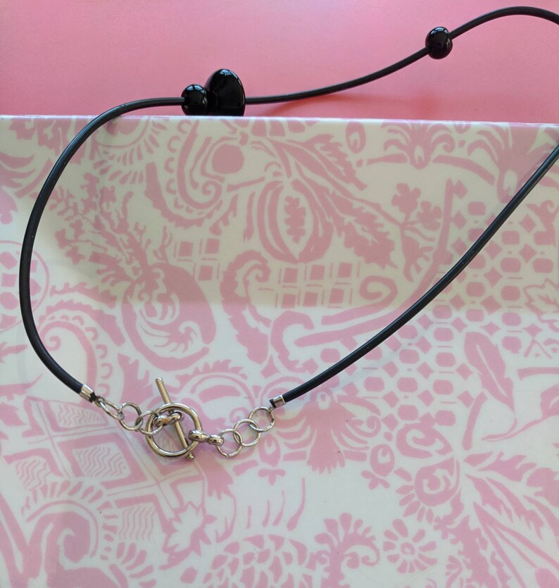 May include: A black cord necklace featuring a silver-toned toggle clasp with a circular ring and a T-bar. The cord is accented with two smooth, round black beads positioned along the length of the necklace, providing a simple and functional design.