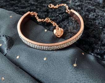 May include: A rose gold-toned bangle bracelet featuring a row of clear, sparkling crystals embedded along the outer surface. A delicate chain is attached to the bangle, ending in a small, polished rose gold-toned heart-shaped charm pendant.