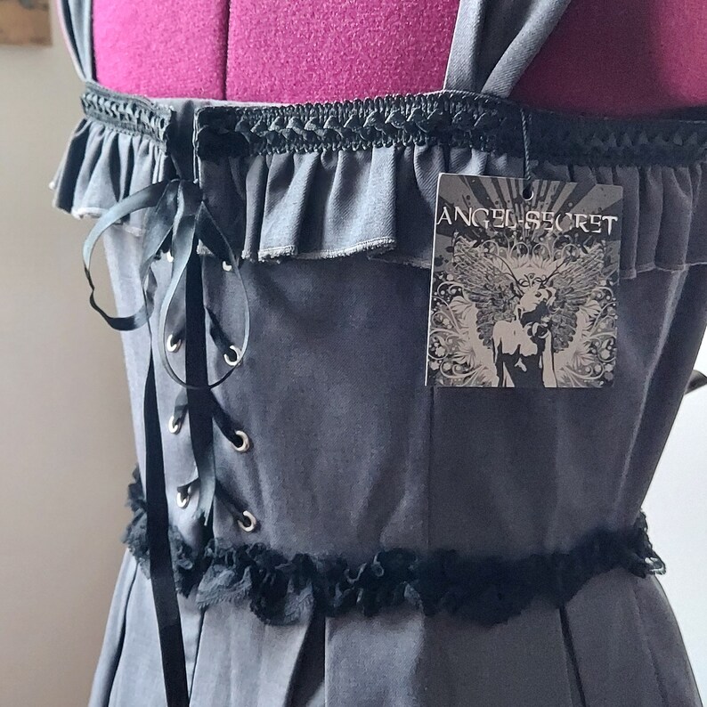 Op de afbeelding: Een grijze jurk met zwarte kantversiering en een zwarte strik om te binden aan de achterkant. De jurk heeft een label met de tekst "Angel Secret".