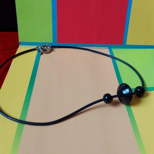 May include: A black cord necklace featuring a central large, flat, circular black bead flanked by two smaller spherical black beads. The necklace is secured with a silver-toned metal toggle clasp, providing a simple and functional design for this piece of jewelry.