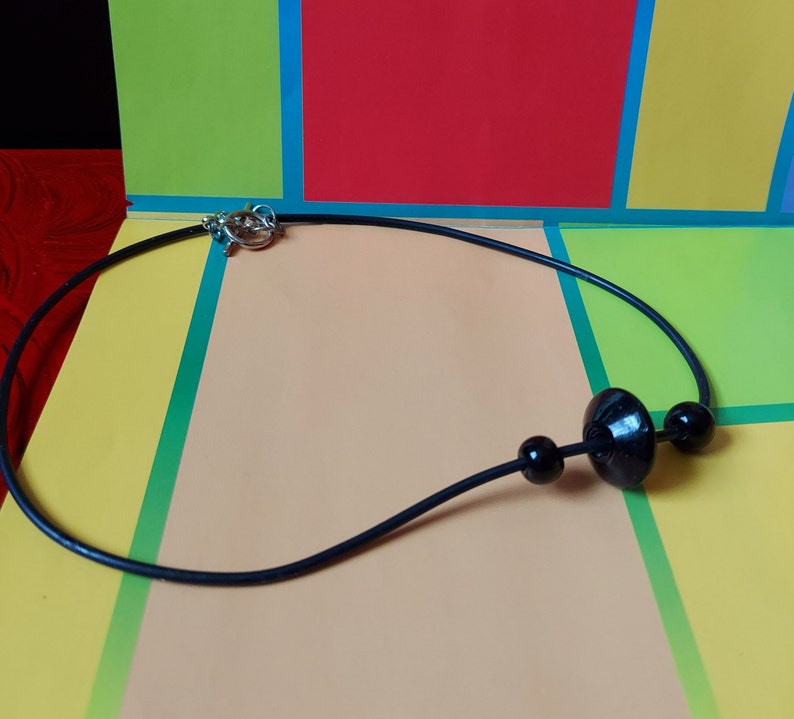 May include: A black cord necklace featuring a central large, flat, circular black bead flanked by two smaller spherical black beads. The necklace is secured with a silver-toned metal toggle clasp, providing a simple and functional design for this piece of jewelry.