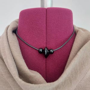 A black cord necklace featuring a central arrangement of three black beads. The design includes two small spherical beads flanking a larger, diamond-shaped bead in the center, all strung on a smooth black rubber cord that encircles the neck. Displayed on a plum color dress form wearing a tan cowl neck dress.