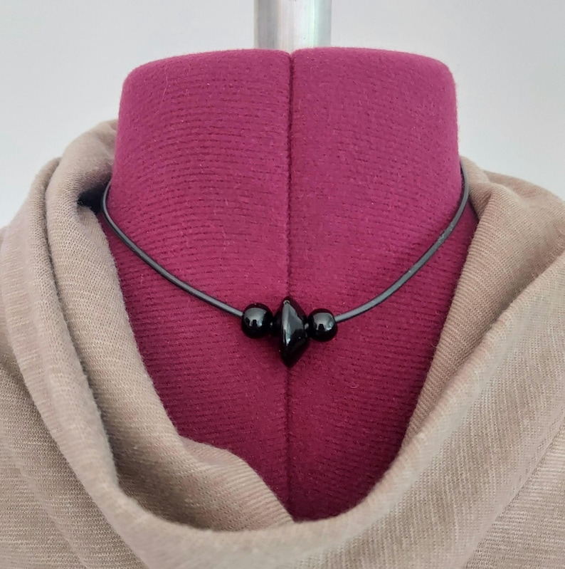 A black cord necklace featuring a central arrangement of three black beads. The design includes two small spherical beads flanking a larger, diamond-shaped bead in the center, all strung on a smooth black rubber cord that encircles the neck. Displayed on a plum color dress form wearing a tan cowl neck dress.