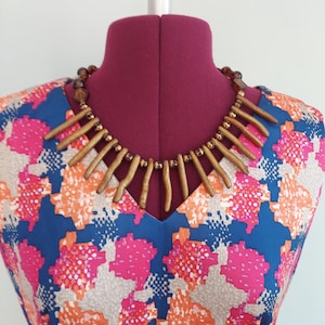 May include: A statement necklace featuring a series of elongated, matte gold-toned cylindrical beads arranged in a fringe pattern, interspersed with smaller, dark brown faceted beads on a dark brown cord, displayed on a dress form.