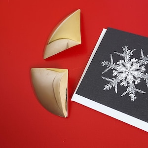 May include: Two gold-toned, curved metal objects and a black and white card with a snowflake design are displayed on a red surface. The metal objects have a rounded, triangular shape. The card features a detailed snowflake illustration.