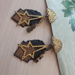 May include: A pair of earrings featuring a textured dark brown and black base with a gold-toned hammered star overlay. Each earring includes a semi-circular gold-toned top piece with a radiating pattern and a small dangling metallic key charm.