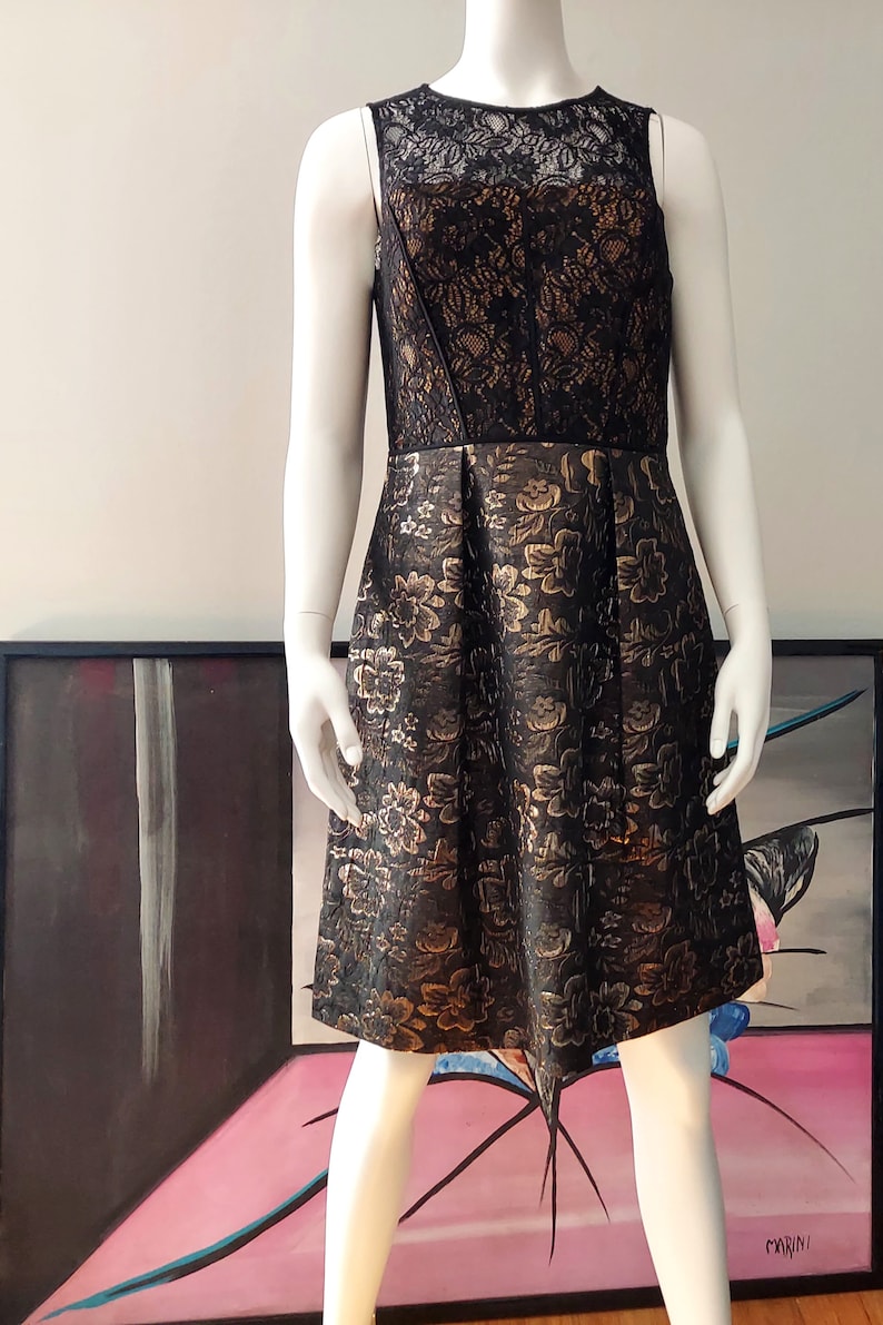 May include: Sleeveless black dress with a floral pattern in gold. The bodice is black lace, and the skirt is pleated. The dress is displayed on a mannequin. The dress is knee-length.