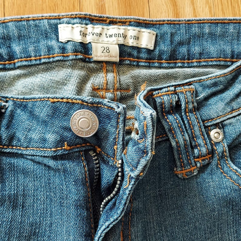 May include: Close-up of blue denim jeans with visible stitching and a metal button. The jeans have a zipper and a label that reads "Forever Twenty One" and size 28.