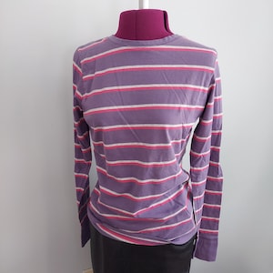 Puede incluir: Una camiseta de manga larga y cuello redondo con rayas horizontales. La camiseta presenta rayas alternas en morado, blanco y rosa. La prenda se exhibe en un maniquí.