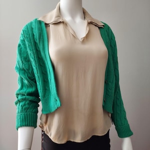Puede incluir: Un cárdigan verde tejido con trenzas, con mangas largas y botones. El cárdigan se lleva sobre una blusa beige con cuello. El maniquí lleva pantalones negros. El look general es casual y elegante.