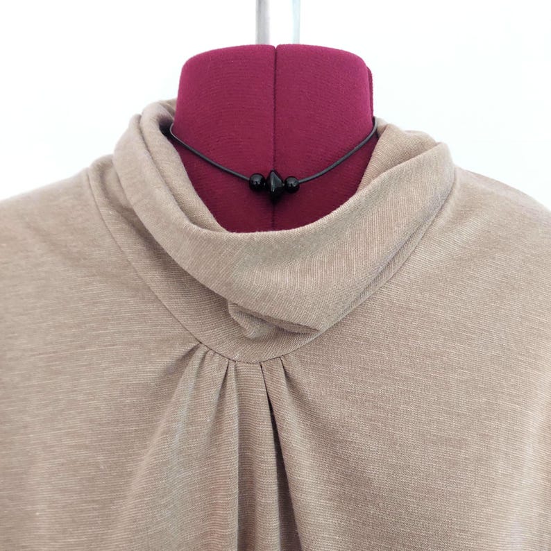 May include: A beige long-sleeve top featuring a draped cowl neckline with subtle pleating at the center front. A thin black cord necklace with three small, smooth black beads is positioned around the neck of the garment on a mannequin.