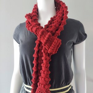 May include: A red knitted scarf with a textured, bobbly design. The scarf is worn around the neck of a mannequin.
