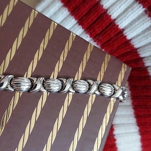 Puede incluir: Una pulsera plateada con un diseño de X y O descansa sobre una caja marrón con rayas diagonales doradas. El fondo presenta un patrón de rayas rojas y blancas.